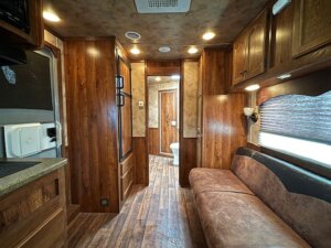 Interior of an RV with wood-paneled walls and floors, a brown sofa on the right, cabinets above, a kitchenette on the left, and a view of a small bathroom—perfect for those seeking Living Quarters custom options or browsing new and used trailers. -Stillwater Trailer Sales