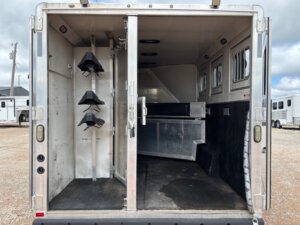 Interior view of a horse and livestock trailer sales unit with open doors, showing saddle racks left and stalls right. The empty trailer, ideal for New and used trailers buyers or Living Quarters custom seekers, is parked on gravel under a cloudy sky. -Stillwater Trailer Sales