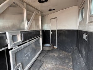 Interior of an empty horse trailer with metal dividers, black and white walls, two small windows, and a spare tire resting on the worn floor. Perfect for those browsing new and used trailers or seeking quality stock trailers for their needs. -Stillwater Trailer Sales