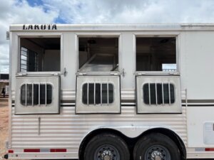 A silver Lakota trailer, ideal for horse and livestock trailer sales, features three open windows with metal bars. Two wheels are visible at the bottom as it rests on a dirt surface beneath a cloudy sky. -Stillwater Trailer Sales