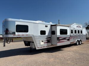 A large white gooseneck horse trailer with a living quarters extension and bold red and black flame graphics, perfect for horse and livestock trailer sales, parked on a gravel lot under a clear blue sky. -Stillwater Trailer Sales