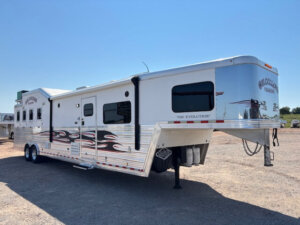 A large silver and white gooseneck horse trailer with black and red flame decals, several windows, and The Evolution on the side, features Living Quarters custom design, parked on gravel under a clear blue sky. -Stillwater Trailer Sales