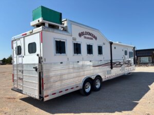 A large silver Bloomer horse trailer with three windows, dual axles, and a green unit on the roof is parked on a gravel lot under a clear blue sky—perfect for those seeking new and used trailers or horse and livestock trailer sales. -Stillwater Trailer Sales