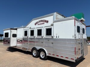 A large white and silver Bloomer brand horse trailer with multiple windows, Living Quarters custom slide-out, parked on gravel under a clear blue sky. -Stillwater Trailer Sales
