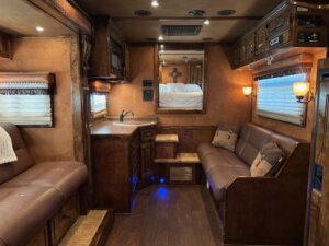 Interior of a luxury RV or trailer, often found in premium stock trailers, featuring brown leather sofas, wood cabinetry, a small kitchen sink, built-in lighting, and a raised sleeping area with a cross decoration on the back wall. -Stillwater Trailer Sales