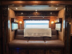 A brown leather sofa with a white blanket draped over the back is set against a window with closed blinds, creating a cozy touch in these Living Quarters custom spaces, often found in premium horse and livestock trailer sales. -Stillwater Trailer Sales