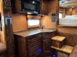 A compact RV kitchen with dark wooden cabinets, a small sink, and a microwave above, showcases Living Quarters custom features. Carpeted steps lead to a raised sleeping area and a window with valance sits above the sink—perfect for trailer sales displays. -Stillwater Trailer Sales
