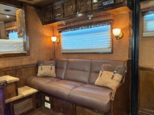A brown leather couch with two pillows sits against a wall with wood paneling and cabinets above—perfect for relaxing after browsing horse and livestock trailer sales. Two wall-mounted lights, a window, and nearby steps complete the inviting space. -Stillwater Trailer Sales