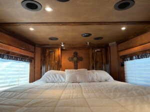A neatly made bed with white bedding sits between two windows under a wooden ceiling with recessed lights; a cross decorates the wood-paneled wall, echoing the rustic charm often found in spaces designed for horse and livestock trailer sales. -Stillwater Trailer Sales