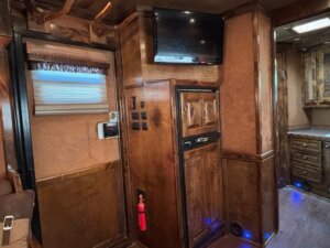 Interior view of a room with wood paneling, a mounted TV above a built-in cabinet, a window with a shade, and kitchen area—showcasing the quality Living Quarters custom options available in our new and used trailers. -Stillwater Trailer Sales