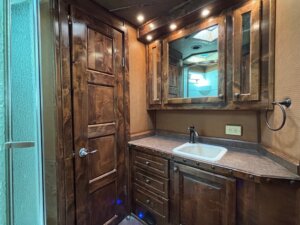 A small bathroom in a Custom Show Trailers unit features dark wood cabinets and a matching door, a countertop with sink, wall-mounted mirror with lights above, towel ring, electrical outlet, and a shower stall visible on the left. -Stillwater Trailer Sales