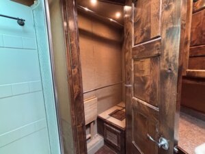 A small, enclosed room with wood paneling and a built-in bench next to a closed wooden door; similar finishes are often found in Living Quarters custom options for new and used trailers. A shower with white tiles is partially visible to the left. -Stillwater Trailer Sales