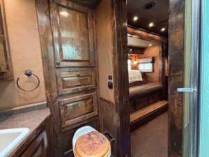 An RV interior features a small bathroom with a wooden toilet seat, towel ring, and cabinets. The bathroom opens to Living Quarters custom-designed for comfort, with a brown couch, wooden accents, and a window with blinds. -Stillwater Trailer Sales