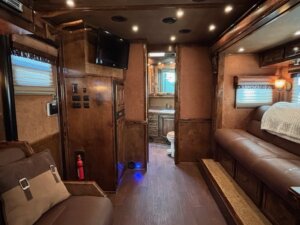 Interior of a motorhome with brown leather seating, wooden cabinets, a mounted TV, and a fire extinguisher—perfect for road trips. Peek into the small bathroom. See more like this among our new and used trailers at our trailer sales showroom. -Stillwater Trailer Sales