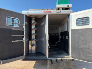 The image shows the open back doors of a horse and livestock trailer. One side features vertical racks and hooks for tack storage, while the other side is an empty stall for horses. The ground is dirt and the sky is clear and blue. -Stillwater Trailer Sales