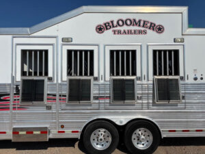 A silver Bloomer Trailers horse trailer with three barred windows and lower vents, parked on gravel. This Stock Trailer features a logo and rides on two visible wheels, perfect for those needing reliable livestock transport. -Stillwater Trailer Sales