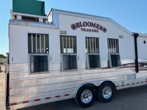 A white Bloomer Trailers horse trailer, ideal for Custom Show Trailers or Stock Trailers needs, with three barred windows and dual wheels, is parked on a gravel lot under a clear blue sky. -Stillwater Trailer Sales