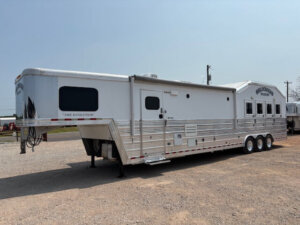 A large silver horse trailer with three axles and multiple windows is parked on gravel. The words The Evolution and 4 Star Trailers are visible on the side and front. The background shows an open outdoor area. -Stillwater Trailer Sales