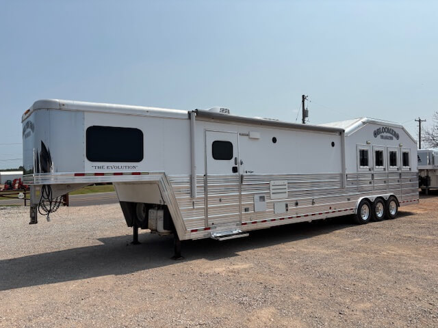A large, silver gooseneck horse trailer with three axles and multiple windows is parked on gravel. The trailer has “THE EVOLUTION” and “SUNDOWNER” branding visible on its exterior. -Stillwater Trailer Sales