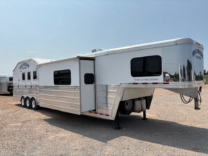 A large, silver gooseneck horse trailer with multiple windows, three axles, and the words THE EVOLUTION on the front, parked on a gravel lot under a clear sky. -Stillwater Trailer Sales