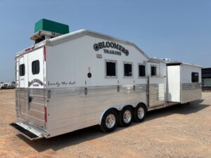 A large, silver Bloomer trailer with three axles is parked on a dirt lot. The trailer has multiple windows, a side slide-out, and a green unit mounted on the roof. The text Only the best is visible on the side. -Stillwater Trailer Sales
