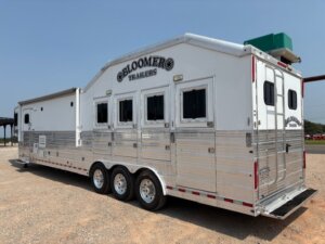 A large white and silver Bloomer horse trailer with three axles, several windows, and a vent on the roof, parked on a gravel lot under a clear sky. -Stillwater Trailer Sales