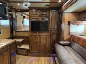 A living area inside an RV with wood cabinets, a flat-screen TV, a brown sofa, carpeted steps leading to a raised sleeping area, wood floors, and windows with blinds and valances. -Stillwater Trailer Sales