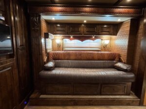 A large brown leather sofa with wooden armrests and storage drawers underneath sits against a wall with wood-paneled cabinets, small lights, and a window with shades in a room with dark wood decor. -Stillwater Trailer Sales