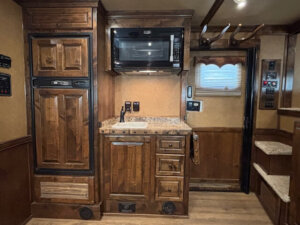A small kitchen area with wood cabinetry, a refrigerator on the left, a microwave above a marble-patterned countertop with a sink below, drawers, and a door with a window on the right. Stairs are visible in the corner. -Stillwater Trailer Sales