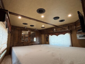 A spacious RV bedroom features a large bed with a white mattress, wood-paneled walls and cabinets, two windows with blinds, ceiling lights, air vents, and a wall-mounted TV in the corner. -Stillwater Trailer Sales