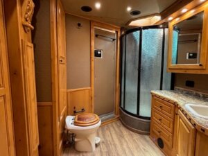 A bathroom with wood-paneled walls and cabinetry, a toilet with a wooden seat, a curved glass shower enclosure, a countertop with a sink, and a mirrored cabinet. The floor has a wood-like finish. -Stillwater Trailer Sales