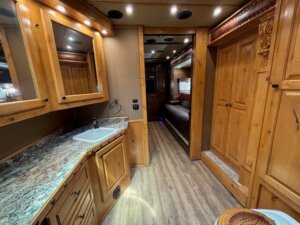 A well-lit RV interior with wood cabinetry, a marble-look countertop with a sink, a wood-paneled refrigerator, overhead lights, wood laminate flooring, and a hallway leading to a bunk bed and a window in the background. -Stillwater Trailer Sales