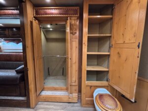 A wooden-paneled bathroom with an open shower stall, shelves with cabinet doors, and a toilet with a wicker-style seat. A sofa is partially visible on the left side of the image. -Stillwater Trailer Sales