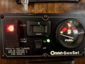 Close-up of an Onan GenSet generator control panel showing indicator lights, an hour meter reading 11, an illuminated red warning light, a fuel gauge just below full, and a switch in the ON position. -Stillwater Trailer Sales
