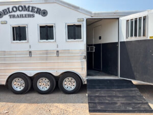 A white Bloomer horse trailer with three axles is shown with its side door open and a black ramp extended to the ground. Several small windows are visible along the trailer. -Stillwater Trailer Sales