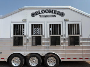 A silver Bloomer horse trailer with three axles, four barred windows with drop-down panels, and the brand name Bloomer Trailers displayed near the roof. -Stillwater Trailer Sales