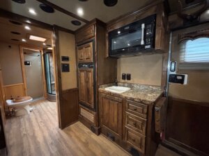 Interior of an RV or camper with a small kitchen featuring a sink, microwave, stove, wood cabinets, and a marble-patterned countertop; a bathroom with shower and toilet is visible in the background. -Stillwater Trailer Sales
