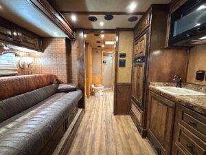 A well-lit RV interior with wood-paneled cabinets, a brown leather sofa on the left, a small kitchenette on the right, and a bathroom with a visible toilet at the end of a hallway. -Stillwater Trailer Sales