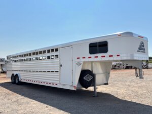 An Elite 32 Combo large, silver gooseneck livestock trailer with side vents and dual axles is parked on a gravel lot under a clear blue sky. The trailer has a covered front hitch, with several other trailers visible in the background. -Stillwater Trailer Sales