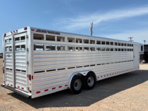 A large, silver Elite 32' Combo livestock trailer with slatted sides and two axles is parked on a gravel lot under a clear sky. The trailer appears empty and is designed for transporting animals. -Stillwater Trailer Sales