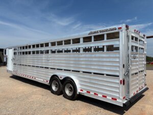 An Elite 32 Combo, large silver livestock trailer with dual rear axles, is parked on a gravel surface under a blue sky. The trailer has slatted sides for ventilation and red and white reflective tape along the bottom edges. -Stillwater Trailer Sales