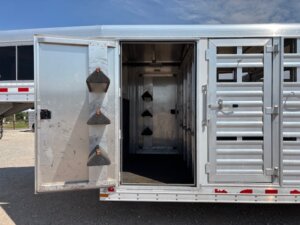 An open metal Elite 32 Combo livestock trailer with the side door open, showing angled black saddles on the door and an empty interior featuring several saddle racks mounted on the walls. -Stillwater Trailer Sales