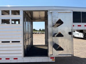 A silver Elite 32 Combo livestock trailer with an open side door reveals an interior with saddle racks on the door and open stalls; trucks are visible in the background under a clear blue sky. -Stillwater Trailer Sales