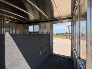Interior of an Elite 32 Combo metal livestock trailer featuring a carpeted partition wall, open door revealing a gravel lot and sky outside, and sturdy metal hooks on the right wall. -Stillwater Trailer Sales