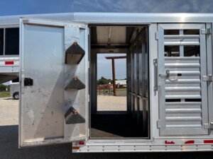 Open door of an Elite 32 Combo silver livestock trailer reveals an interior with saddle racks on the left and divided compartments inside; a farm setting is visible in the background through the trailer. -Stillwater Trailer Sales