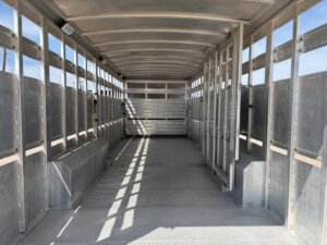 Interior view of an empty Elite 32 Combo metal livestock trailer with a corrugated floor, open sides, and divided sections. Sunlight streams through the slats, casting shadows on the floor and walls. -Stillwater Trailer Sales