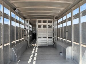 The interior of an empty Elite 32' Combo livestock trailer features metal walls, grated windows, and a divider gate near the front; sunlight casts shadows on the floor. -Stillwater Trailer Sales