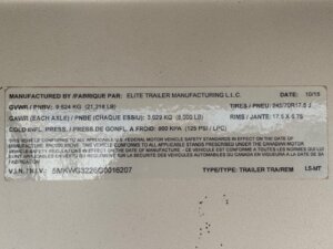 A metal label displays manufacturing information for an Elite 32 Combo Trailer, including weights, tire and rim specifications, VIN, and compliance details in English and French. The label appears slightly worn. -Stillwater Trailer Sales