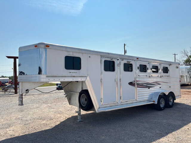 A Dream Coach 4 horse trailer with four windows and a graphic design is parked on a gravel lot under a clear blue sky. The silver gooseneck horse trailer features two axles and a raised front section. -Stillwater Trailer Sales