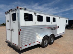 A white Dream Coach 4 Horse trailer with four windows and decorative decals is parked on a gravel lot under a blue sky. The gooseneck horse trailer features two axles and ventilation slats along the sides. -Stillwater Trailer Sales